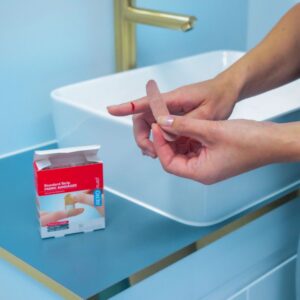 Person applying an AeroPlast fabric bandaid to a finger at a bathroom basin, with the box of 100 beside the sink